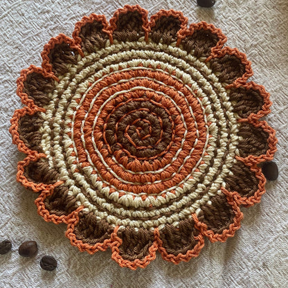 Round crochet mat with concentric circle pattern in brown, orange, and beige on a textured surface.