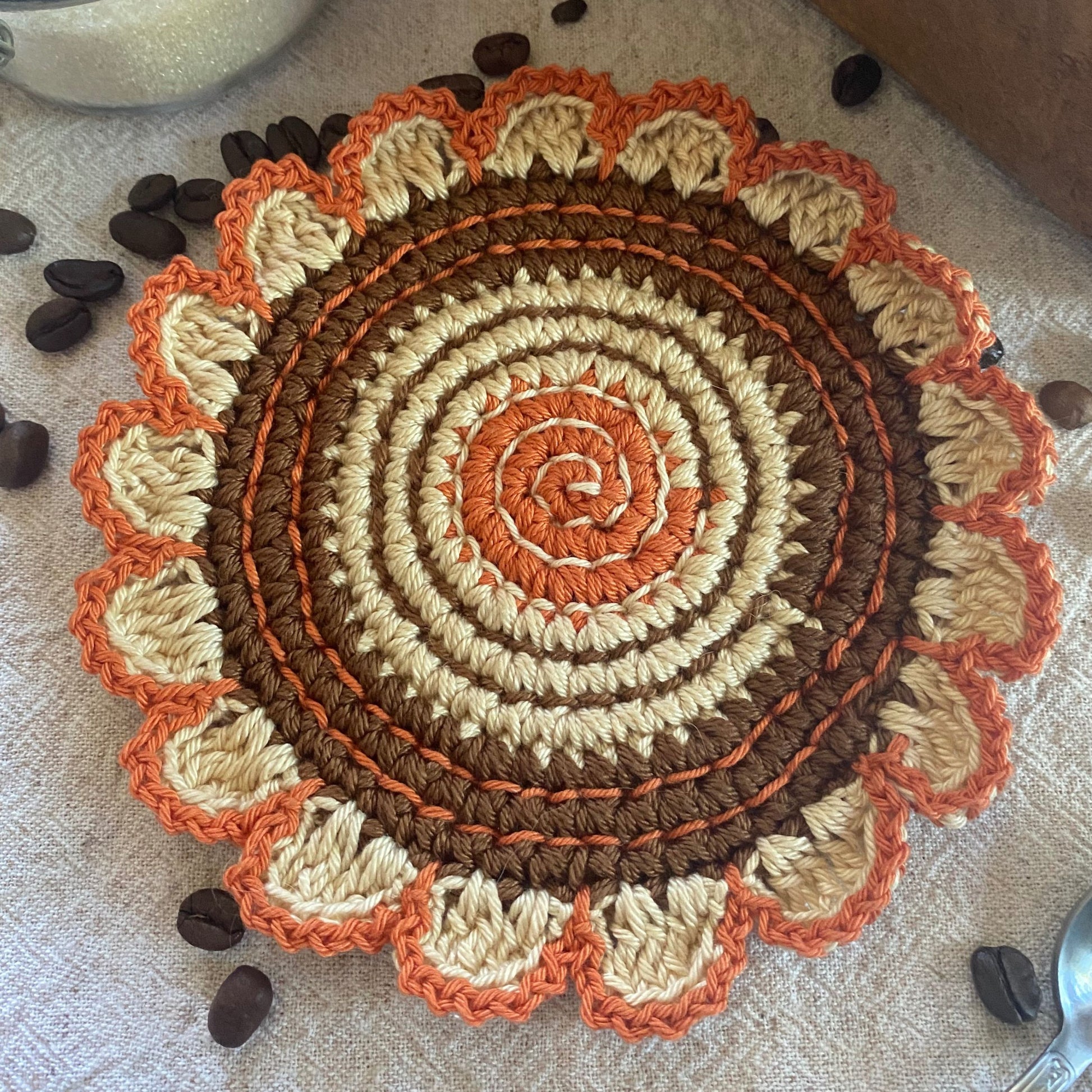 Round crochet trivet with spiral pattern in brown, orange, and beige on a textured surface with coffee beans.