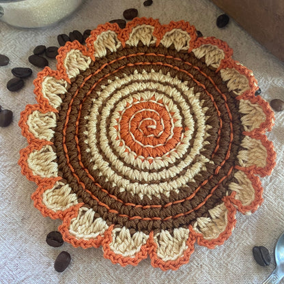 Round crochet trivet with spiral pattern in brown, orange, and beige on a textured surface with coffee beans.