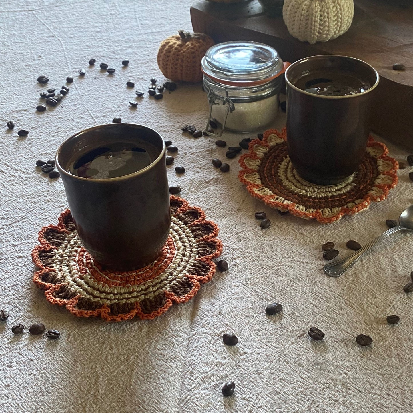 Two cups of coffee on crocheted coasters with scattered coffee beans and a jar on a textured surface.