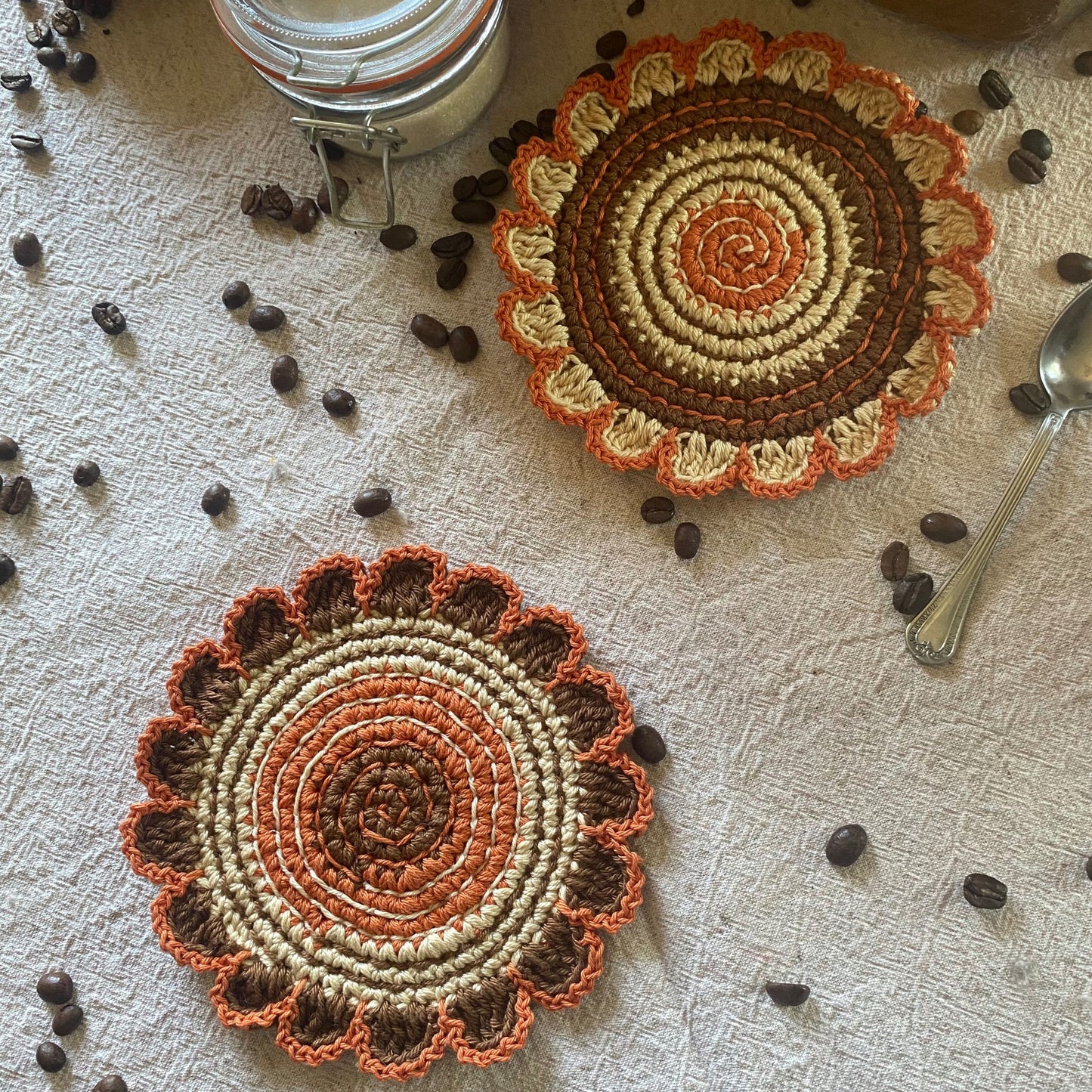 Two crocheted coasters with a floral pattern on a textured surface with coffee beans and a spoon.