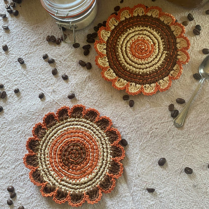 Two crocheted coasters with a floral pattern on a textured surface with coffee beans and a spoon.