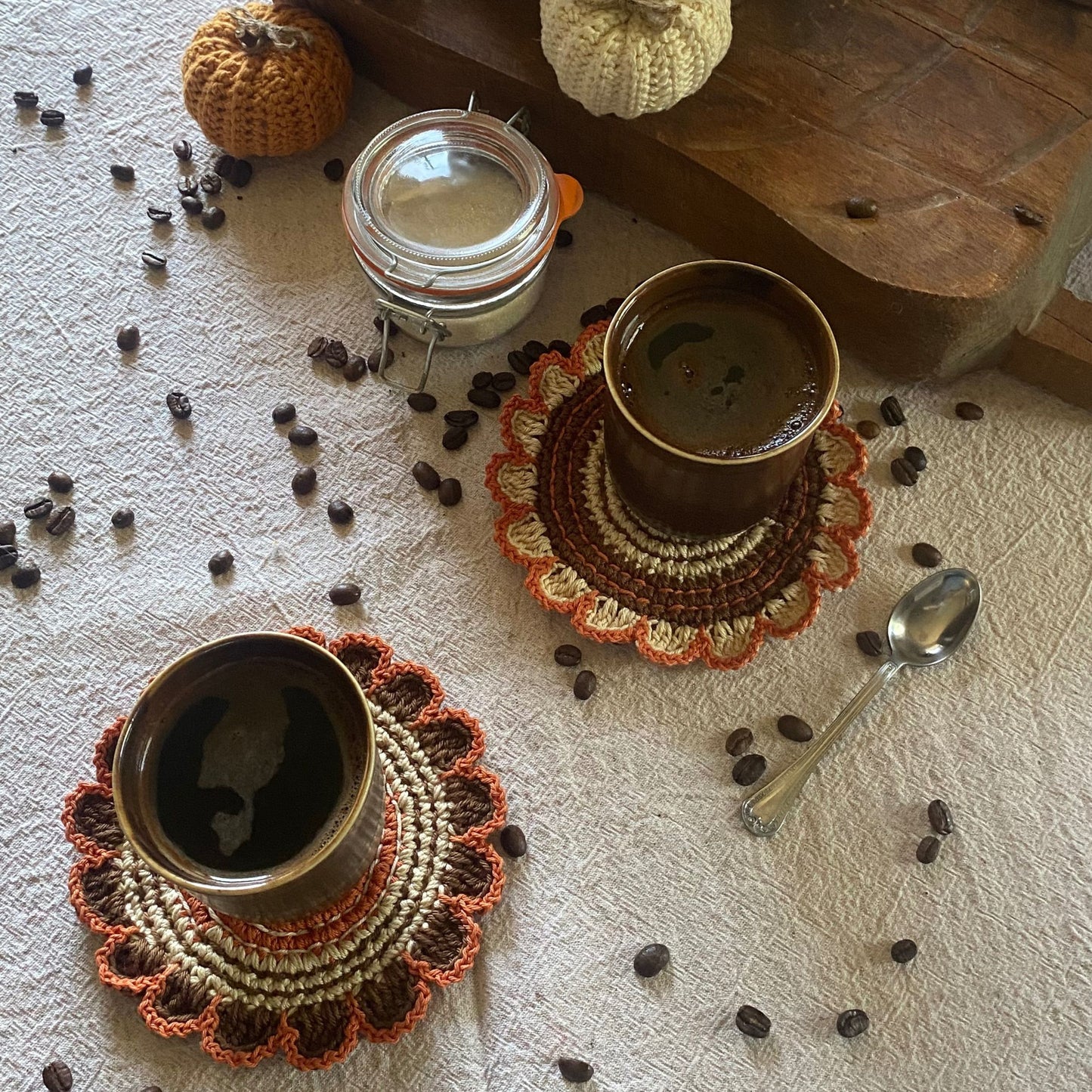 Two cups of coffee on crochet coasters with scattered coffee beans and a jar on a textured surface.