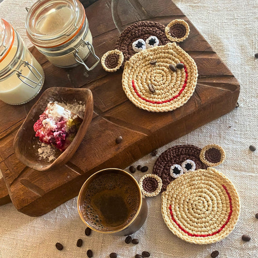 Top-down view of two handmade crochet monkey coasters made of 100% cotton on a wooden board next to a cup of coffee.